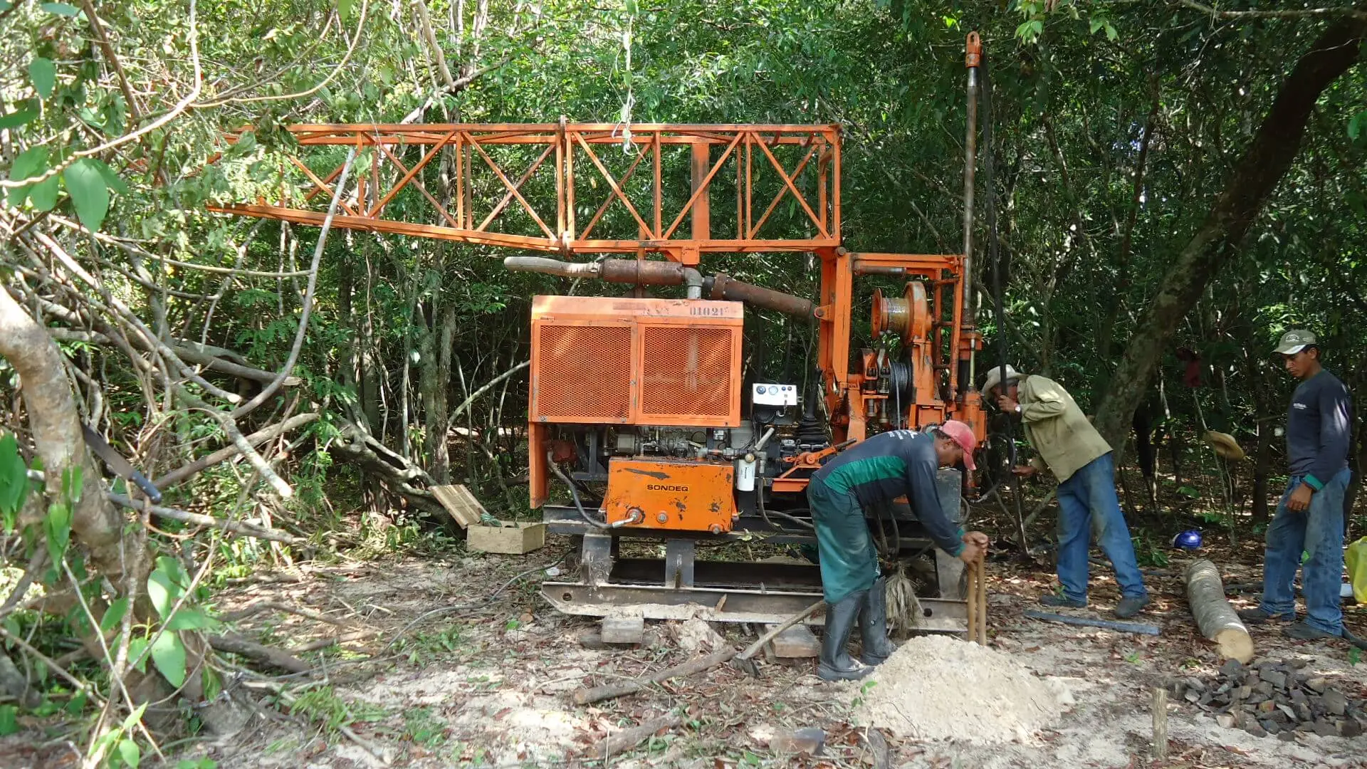 Sonda rotativa laranja operando em clareira de mata fechada, com três profissionais da Geosul manobrando a coluna de perfuração